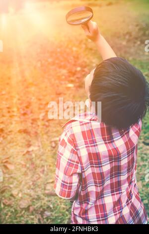 Glückliches asiatisches Kind genießt in der Natur. Kleiner Junge, der die Natur erkundet und durch eine Lupe nach oben schaut. Tagsüber im Freien mit hellen Sonnenauf Stockfoto