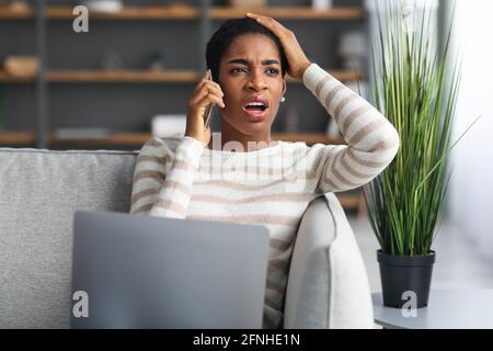 Problem Mit Dem Internet. Verwirrt Schwarze Frau Im Gespräch Auf Handy Und Mit Laptop Stockfoto