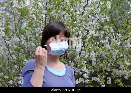 Frau, die medizinische Gesichtsmaske in einem Frühlingsgarten auf Kirschblütengrund entfernt. Konzept des Genußens der Blumen Geruch, Ende der Quarantäne Stockfoto