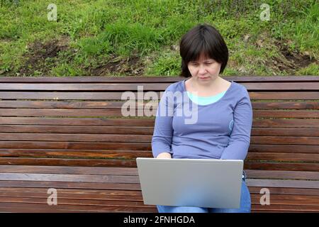 Frau, die mit einem Laptop auf ihrem Schoß auf einer Holzbank im Park sitzt. Konzept für Remote-Arbeiten im Freien, freiberuflicher Mitarbeiter Stockfoto