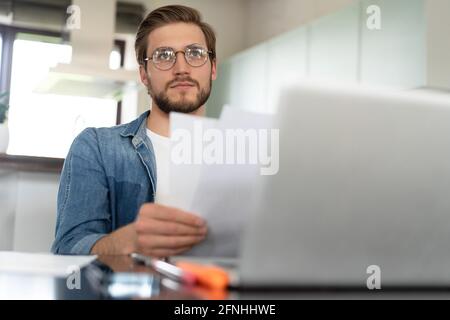 Junger Mann, der von zu Hause aus mit einem Laptop arbeitet und Papierkram macht. Stockfoto