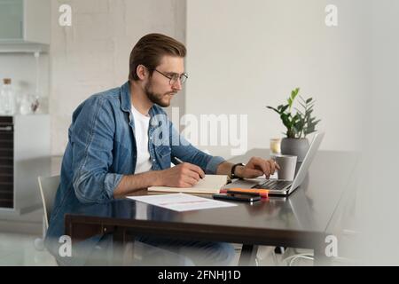 Junger Mann, der von zu Hause aus mit einem Laptop arbeitet und Papierkram macht. Stockfoto
