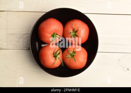 Mehrere reife rosafarbene Tomaten in einer schwarzen Keramikplatte, Nahaufnahme, auf einem weißen Holztisch, Draufsicht. Stockfoto