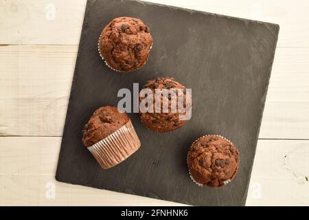 Vier Schokoladenmuffins auf einem Schieferbrett, auf einem weißen Holztisch, Draufsicht. Stockfoto