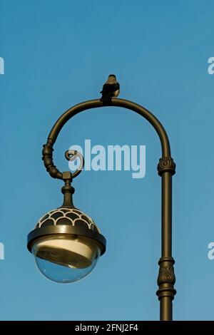 Fragment einer Straßenlaterne der Stadt, auf der eine Krähe gegen einen blauen Himmel sitzt. Stockfoto