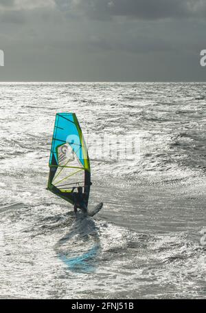 Auffallendes Bild eines einzelnen Windsurfers mit blauem Segeltörn Zusammen mit einem dunklen Himmel Hintergrund vor einer silbernen gesetzt Meer Stockfoto