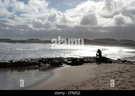 Eine Figur in Silhouette, sitzend auf Felsen, denkend und in Ruhe, während man auf das Meer blickt, mit einem Hintergrund aus Dünen und treibenden Wolken Stockfoto