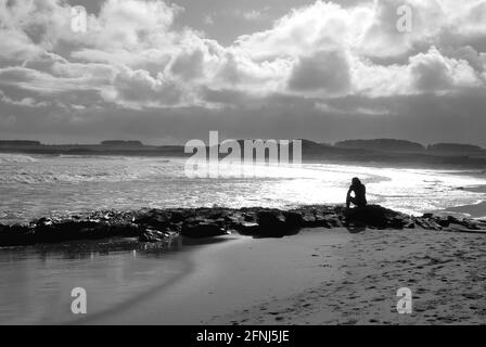 Ein Monobild einer einzelnen Figur in sitzender Silhouette Auf Felsen an der Küste und mit Blick aufs Meer Mit Dünen und treibenden Wolken als Hintergrund Stockfoto