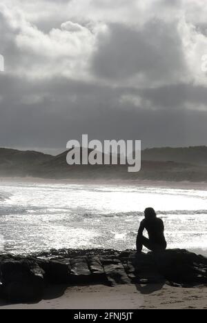 Ein Bild im Hochformat einer einzelnen Figur leise Sitzen auf Felsen in einer Küstenlandschaft mit einer silbrigen Meer und Hintergrund von Dünen und Wolken Stockfoto