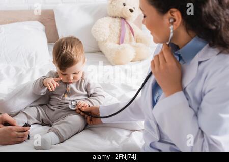 Verschwommener afroamerikanischer Kinderarzt im weißen Mantel, der einen Jungen untersucht Mit Stethoskop in der Nähe der Mutter Stockfoto