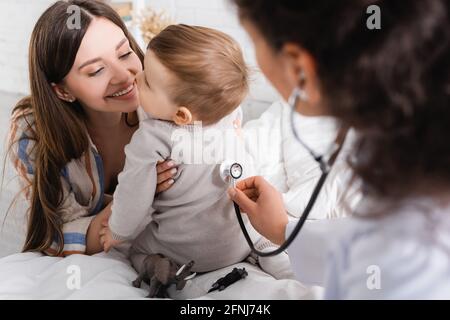 Verschwommener afroamerikanischer Kinderarzt im weißen Mantel, der einen Jungen untersucht Mit Stethoskop Stockfoto