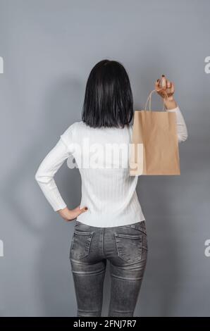 Frau in einem weißen Pullover hält in ihrer Hand eine Bastelpapiertasche zum Einkaufen. Zurück zur Kamera gedreht Stockfoto