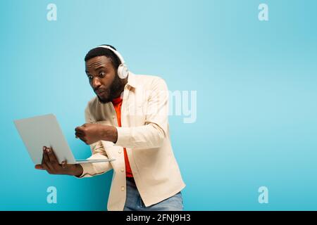 Wütender afroamerikanischer Mann in Kopfhörern, der geballte Faust in der Nähe zeigt Laptop auf blauem Hintergrund Stockfoto