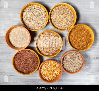 Draufsicht auf verschiedene Vollkorngetreide ( Mais, Reis, Quinoa, Hirse, Roggen, Weizen, Sorghum, Hafer) in runden Keramikschalen auf grauem Holztisch Stockfoto