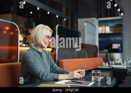 Positive attraktive Frau im Alter in einer Brille, die am Tisch sitzt Modisches Café und Smartphone beim Lesen des Artikels auf dem Handy internet Stockfoto