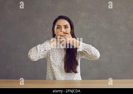 Verängstigte junge Frau, die den Mund mit der Hand bedeckt, die am Tisch sitzt studioaufnahme Stockfoto