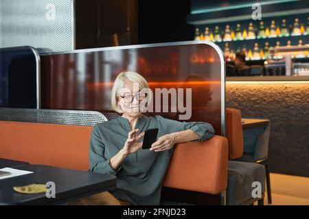 Glückliche schöne ältere Frau mit blonden Haaren auf dem Sofa sitzen Und den Ellenbogen auf dem Rücken des Sitzes anlehnen, während Sie das Smartphone verwenden Für die Online-Kommunikation Stockfoto