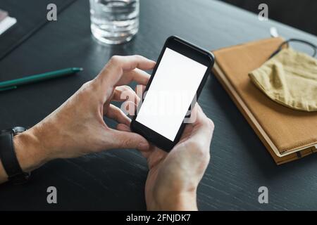 Männliche Hände halten Smartphone mit leerem Bildschirm am Tisch mit Organizer, Stoffmaske und Stift Stockfoto