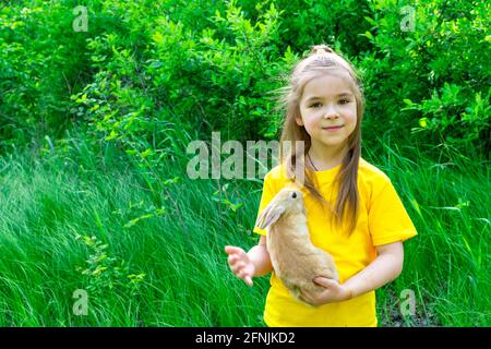 Kleines nettes Mädchen spielt mit einem echten Ingwer-Kaninchen auf einem Hintergrund von grünen Pflanzen. Platz für eine Inschrift. Im Sommer Aktivitäten im Freien für Kinder Stockfoto