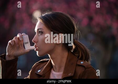 Hübsche junge Frau, die im Frühling vor einem blühenden Baum eine Asthmapumpe benutzt. Saisonaler Allergieanfall Stockfoto