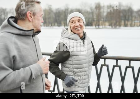 Fröhliche sportliche reife Dame in Handschuhen und Hut lächelnd an Ehemann, während sie gemeinsam am Flussufer im Winterpark entlang laufen Stockfoto