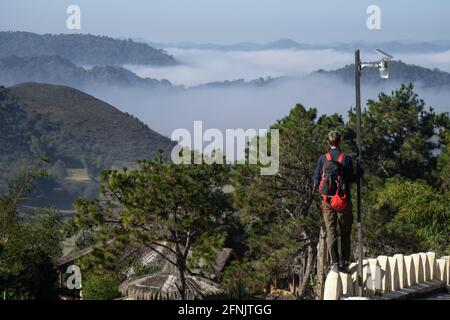 Ein männlicher Toursit mit rotem Rucksack steht auf einem Zaun neben einem Dorf und blickt auf eine neblige Tallandschaft, Shan State, Myanmar Stockfoto