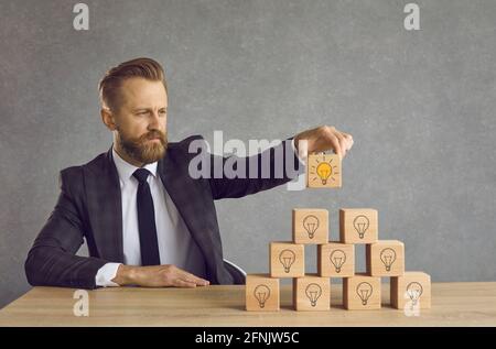 Geschäftsmann findet Lösung und legt den letzten Würfel mit Glühbirne Oben auf seiner Pyramide der Ideen Stockfoto