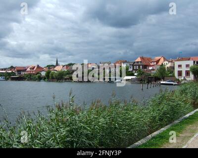 Blick über einige Schilfpflanzen zu einigen Wohnhäusern am See an der Mecklenburgischen Seenplatte. Einige Gewitterwolken brauen über dem Dorf Stockfoto