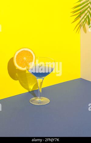 Blauer Cocktail mit einer Scheibe Orange am Rand des Glases auf einer Kombination aus blauem Gelb und cremefarbenem Hintergrund. Palmenblatt auf dem Hintergrund. Summe Stockfoto