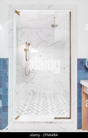 Blick in die Tür einer wunderschönen gefliesten Dusche mit Marmorfliesen, einer Glastür und einem goldenen Duschkopf. Das Badezimmer ist mit blauen Fliesen ausgelegt. Stockfoto