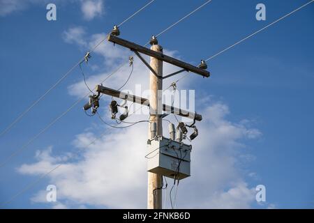 Ein hölzerner Strommast und Transformator mit Kommunikations- und Übertragungsleitungen unter einem blauen Himmel Hintergrund. Stockfoto