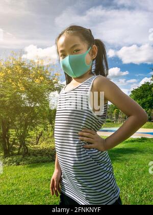 Portrait Asiatische kleine Mädchen posiert im Park mit Arm Akimbo, trägt Schutzmaske für Corona-Virus-Epidemie, Hintergrund blauen Himmel und Blumenbäume, ve Stockfoto