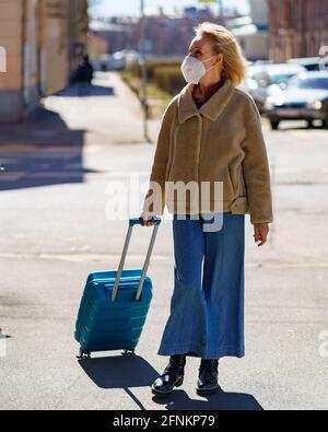 Modische ältere Frau, die in einem Atemschutzgerät oder einer Gesichtsmaske auf einer sonnigen Straße mit blauem Koffer steht und nach oben schaut. Ältere Dame verirrt sich während der Reise während des covid19-Ausbruchs in der Stadt Stockfoto