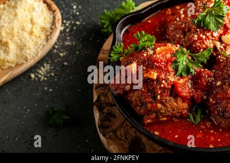 Nahaufnahme von drei traditionellen großen italienischen Fleischbällchen in zischender Tomatensauce in gusseiserner Pfanne. Die Pfanne sitzt auf Holz-Untersetzer und Petersilienblättern Stockfoto