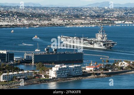 Ein Flugzeugträger, der von einem Schlepper nach San geführt wird Diego Bay in Kalifornien mit einem Marinestützpunkt in der Vordergrund und die Stadt San Diego Stockfoto