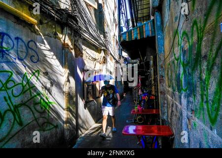 Ein Mann, der einen blauen Regenschirm hält, geht eine sonnendurchflutete Gasse in Chinatown, Bangkok, Thailand, entlang Stockfoto