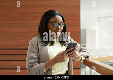 Afrikanische Frau in einer Brille, die ihr Mobiltelefon im Stehen benutzt Auf dem Balkon im Büro Stockfoto
