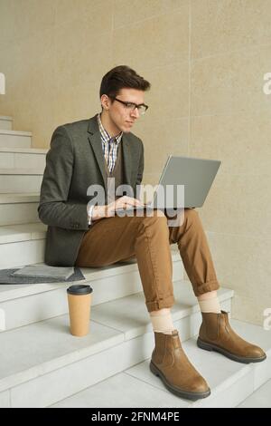 Junger Geschäftsmann in Brillen, der auf der Treppe mit einem Laptop sitzt Knie und Online-Arbeit Stockfoto