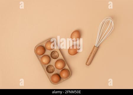 Eier in Holzeischachtel und Schneebesen auf beigem Hintergrund. Flach liegend Draufsicht Kopierbereich. Stockfoto