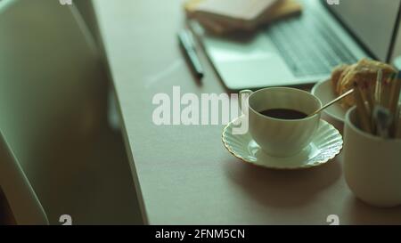 Seitenansicht des Frühstücksmahls mit Kaffeetasse und Croissant Am Arbeitsplatz mit Laptop und Schreibwaren Stockfoto