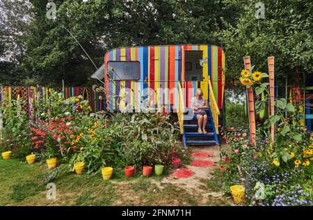 Corps-Nuds, Frankreich. Ille-et-Vilaine Abteilung.. Der bunte Garten bringt einen alten Wohnwagen zusammen, der mit Farblinien bemalt ist, umgeben von sehr c Stockfoto