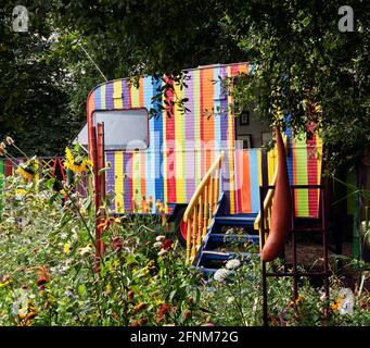 Corps-Nuds, Frankreich. Ille-et-Vilaine Abteilung.. Der bunte Garten bringt einen alten Wohnwagen zusammen, der mit Farblinien bemalt ist, umgeben von sehr c Stockfoto