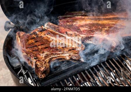 In Knochen gewürzte Rindersteaks Grillen auf einem Grill Feuer sizzling Über den heißen Kohlen mit Rauch in einer Nahaufnahme Blick im Freien Stockfoto