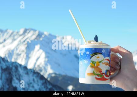 Person, die einen blauen Becher mit dem Bild des Weihnachtsmannes in der Hand hält, der heiße Schokolade mit Schlagsahne in den Europäischen Alpen, Tirol in Österreich enthält Stockfoto