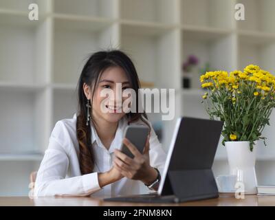 Porträt einer Studentin, die während der Arbeit mit der Kamera in die Kamera schaut Smartphone und Tablet im Café Stockfoto