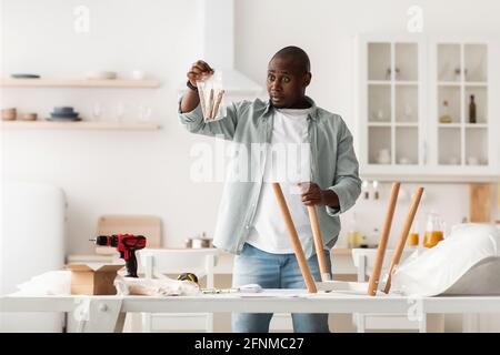 Verwirrt afroamerikanischer Mann hält Paket mit Details, Montage Stuhl mit Anweisungen in der Küche Stockfoto