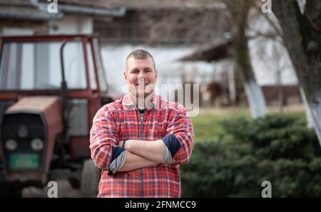 Glücklicher Bauer mit gekreuzten Armen, der vor dem Traktor steht Auf dem Bauernhof Stockfoto