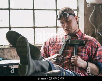 Junger Arbeiter sitzt auf einem Stuhl mit Beinen auf dem Schreibtisch, hält Hammer und Schraubenschlüssel auf der Brust und schläft Stockfoto