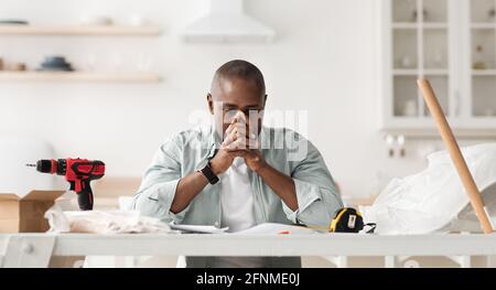 Probleme mit der Möbelmontage. Verstörter schwarzer Mann, der im Kücheninnenraum mit Werkzeugen sitzt und Anweisungen ansieht Stockfoto