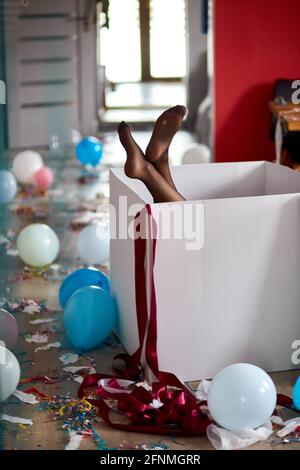 Frau Beine kleben aus großen Box mit undichten Strümpfen, nach Party Chaos, chaotisch im Wohnzimmer zu Hause, Konfetti und Ballons, Morgen nach Party cel Stockfoto